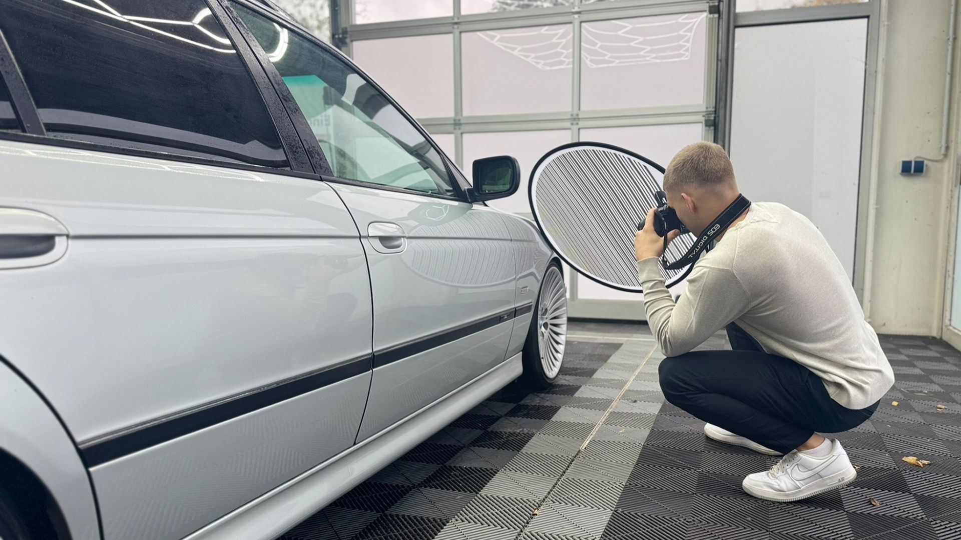 Kfz-Gutachter fotografiert in der Hocke die vordere Kotflügelpartie eines silbernen Autos mithilfe eines runden Reflexionssegels zur Schadensdokumentation